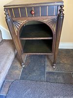 Photo showing table front view with single drawer at top and two open shelves below, carved decorative legs and edges, placed on tiled floor next to carpeted stairs.