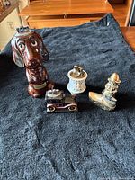 Four table lighters including a brown ceramic dog, white ceramic decorative lighter, vintage car lighter, and metal novelty fisherman lighter arranged on a dark cloth.