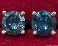 Close-up of the pair of 14KT white gold earrings with round blue diamonds set in four prong settings on a red background showing color and cut detail.