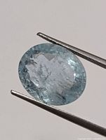 Close-up photo of the oval faceted aquamarine gemstone held by tweezers, showing internal inclusions and light blue color.