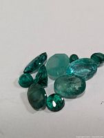 Multiple loose emerald gemstones arranged on white background showing various cuts and shapes with green color and natural inclusions.