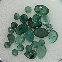 Top-down close-up of 26 genuine emerald gemstones of varying shapes and sizes with visible natural inclusions, laid out on a white surface.