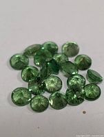 Close-up photo of seventeen small round faceted green gemstones spread on a white surface.