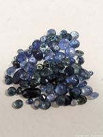Pile of loose genuine sapphire gemstones with various cuts and blue colors displayed on a light surface.