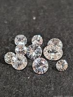 Ten round brilliant cut white sapphire loose stones displayed on a dark fabric background, showing clarity and sparkle.