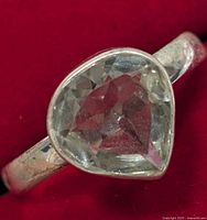 Close-up photo of a silver ring with a pear-shaped green amethyst gemstone showing its cut and clear faceted surface on a red background.