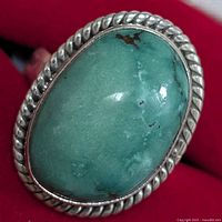 Close-up of oval turquoise cabochon ring with twisted silver rope bezel on red cloth background, showing color and texture of stone.