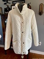 Front view of cream-colored quilted jacket on mannequin with buttons and pockets visible.