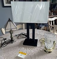 Three lamps on a white crocheted tablecloth: a white stained glass lamp with bronze base, a modern rectangular white fabric shade lamp with black base, and a small ribbed clear glass lamp on brass fittings.