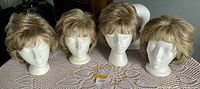 Front view of four light brown wigs placed on three white styrofoam mannequin heads displayed on a crochet tablecloth.