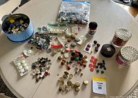 Wide view of multiple bags, tins, and loose buttons spread out on a table showing various button styles and sizes.