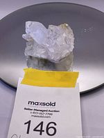 Clear raw quartz crystal cluster shown on reflective surface with auction label in front.