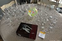 Set of about twenty clear crystal wine glasses, a colorful floral plastic juice jug, and a boxed zinc alloy corkscrew set on a lace tablecloth.