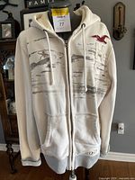 Front view of a white zip-up Hollister hoodie with distressed grey stripes and red logo on chest.