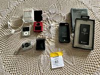 Photo showing all items laid out on a crocheted beige doily with the lot label visible. Items include iPod Nano with headphones, mini digital camera, iPhone charger case with packaging, cables, a camera keychain, and pouch.