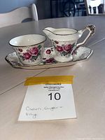 Set of porcelain cream and sugar vessels and tray with vintage rose floral pattern and gold accents shown on a grey table surface.