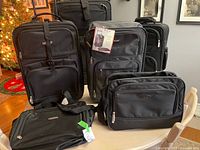 Seven black luggage and travel bags displayed on table including two medium wheeled suitcases, three smaller carry bags, one duffel, and one shoulder bag.