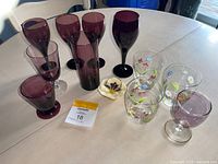 Top view of table with 12 pieces including dark purple goblets, purple tinted glasses, clear hand painted floral glasses, and small flower dish in center.