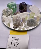 Lot of raw mineral specimens arranged on a mirrored surface showing green fluorite, purple amethyst, quartz, and other crystals.