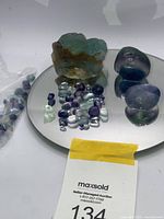 Photo showing a large raw fluorite crystal, multiple small tumbled fluorite stones in green, purple and clear shades, and three larger polished stones displayed on a reflective surface.