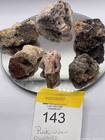 Six raw mineral crystal specimens with pink and grey tones displayed on a mirrored surface with an auction tag.