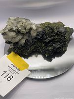 Raw mineral specimen featuring greenish-black epidote and white quartz on dark rock shown on a mirror base to highlight size and detail.