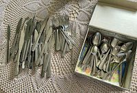 Mixed stainless steel utensils spread out and inside a box on a lace tablecloth showing knives, forks, teaspoons, and soup spoons in differing patterns and styles.