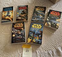 Six Star Wars X-Wing series paperback novels arranged on a crocheted tablecloth with a tags showing '23 Books'. Covers depict various starfighter combat scenes.