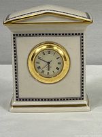 Front view of small white ceramic clock with black decorative trim and circular clock face with Roman numerals in gold bezel.