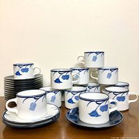 Full set of 13 cups and 14 saucers arranged in stack and grouping, showing Tivoli Belle Fleurs blue floral design on white fine china with one cup on a white saucer and one on a blue saucer.