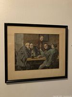 Framed print showing men gathered at table in an indoor setting, muted earthy colors, black frame.