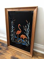 Framed tinsel foil art featuring two orange flamingos with foliage on a black background, displayed on a wooden floor against a white wall.