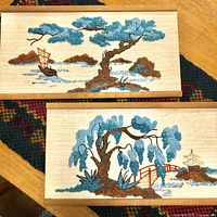 Pair of rectangular gravel art panels with teak edges depicting Asian landscapes with blue trees and traditional elements, placed on woven cloth.