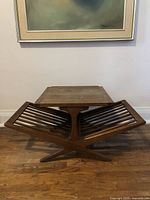 Front view of the mid century modern wooden side table showing the square top and the integrated slatted magazine rack below with cross-legged base on hardwood floor.