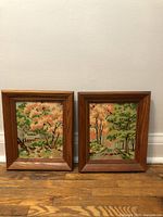 Pair of framed paint by number paintings showing autumn trees with leafy branches in natural settings, side-by-side on floor against white wall.