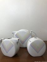Front view of vintage ceramic Toscany tea set items with rainbow chevron design on white background, teapot centered with creamer and sugar bowl flanking it.