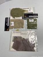 Four unopened packages of camouflage netting fabric from Verlinden and Pegasus, including green, beige, brown, and purple-brown colors.