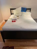 Front view of bed showing dark wood frame, mattress, box spring with protective covers, white bedding, four pillows including blue decorative pillow, and some papers on bed.