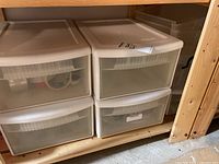 Four white plastic storage drawer units on wooden shelving in basement, contents include adhesive tape and craft supplies.