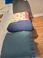 Four blankets stacked on bed: light blue with fringe, dark blue, white with pink pattern and trim, grey
