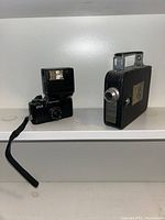 Kodak 8mm movie camera and Pentax Super 110 camera with flash shown side by side on a white shelf. Both cameras are vintage and compact.