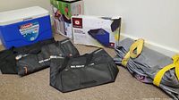 Lot showing blue Coleman cooler, two black insulated cooler bags, gray air mattress bag with yellow straps, and boxed electric air mattress pump.
