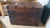 Front view of antique wooden storage trunk chest showing metal corner brackets and side handles. Surface worn with scratches and wear marks.