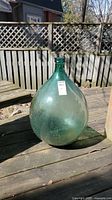 Green glass demi john bottle on wooden surface outdoors, showing full body and narrow neck, surface dirt present.