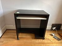 Front view of black desk on wheels with extended white keyboard shelf under table top, placed on hardwood floor against white wall.
