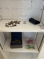 Coins spread on a white surface with two silver coins separated on the right and a dark coin purse on the side; includes a small box and two foreign banknotes below.