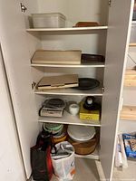 Full view of kitchen cabinet with shelves showing various kitchen items including storage containers, wooden and metal trays, mixing bowls and tools in bags below.