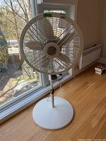 Full view of white Lasko pedestal fan standing on wooden floor by window, showing mesh blade grille and height adjustment mechanism.
