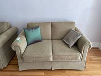Front view of beige upholstered two-seat love seat with two decorative pillows on hardwood floor.