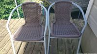 Two patio chairs side by side on a wooden deck with white metal frames and brown woven wicker seats and backs.